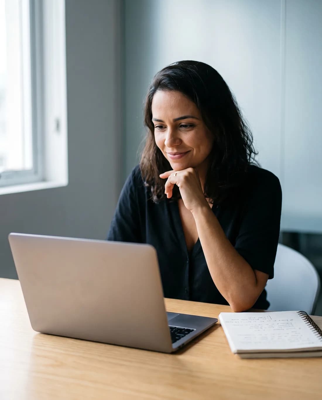 Mulher estudando no notebook, com um caderno de anotações ao lado